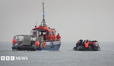 A French rescue boat with crew members in orange uniforms and life jackets escorts a small, overcrowded inflatable boat. The inflatable boat is filled with people wearing life jackets, believed to be migrants, as it moves away from the French coast.