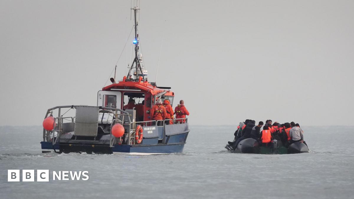 A French rescue boat with crew members in orange uniforms and life jackets escorts a small, overcrowded inflatable boat. The inflatable boat is filled with people wearing life jackets, believed to be migrants, as it moves away from the French coast.