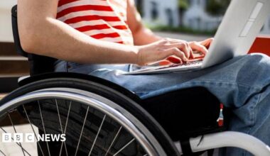 A close-up image of a wheelchair as a woman sits in and works on a laptop. Her jeans and red and white T-shirt and hands and forearms are visible.