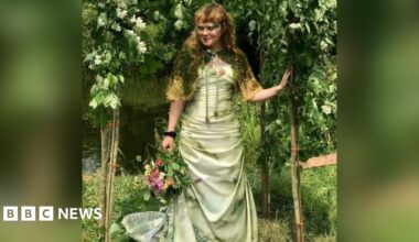 Megan Ruth-Trump stands beneath a floral arch beside a river. She is wearing a wedding dress and holding a bouquet of flowers in one hand