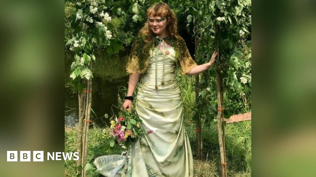 Megan Ruth-Trump stands beneath a floral arch beside a river. She is wearing a wedding dress and holding a bouquet of flowers in one hand