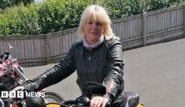 Mel Wallace is a 59-year-old woman with shoulder-length blonde hair. She is sitting astride a yellow motorbike and is holding the handlebars. Mel is wearing a leather jacket and a light scarf around her neck. There are several motorbikes to her right and she seems to be in a car park with a wooden fence behind her, which is in front of a row of bushes