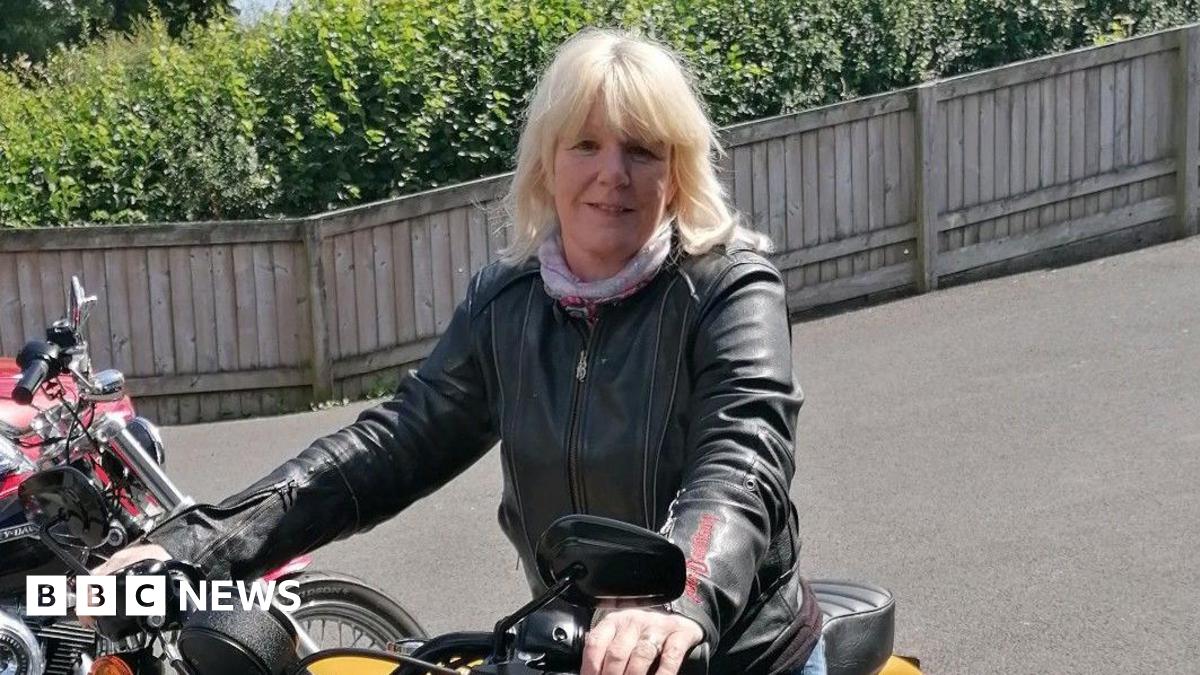 Mel Wallace is a 59-year-old woman with shoulder-length blonde hair. She is sitting astride a yellow motorbike and is holding the handlebars. Mel is wearing a leather jacket and a light scarf around her neck. There are several motorbikes to her right and she seems to be in a car park with a wooden fence behind her, which is in front of a row of bushes