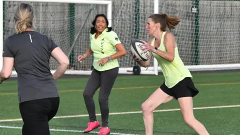 Washington Rugby Club Three women playing touch rugby. A slender woman in the centre has caught the ball midstride. Two other players are running along besides her.
