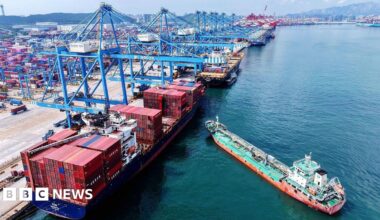 Aerial view of a container terminal in Qingdao with blue cranes hovering over container ships and a smaller green and orange boat approaching in east China's Shandong province Monday, 11 August, 2025.