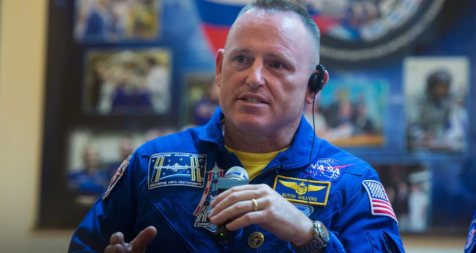 a middle-aged astronaut with a buzz cut and wearing a blue flight suit speaks into a microphone