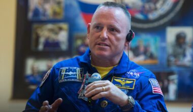 a middle-aged astronaut with a buzz cut and wearing a blue flight suit speaks into a microphone