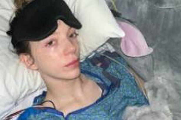 A woman in a blue hospital gown, surrounded with soft toys, remains in bed, with a black eye mask resting on her forehead. 