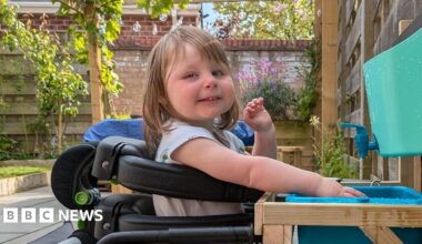 A three-year-old girl in a wheelchair playing with water. She is smiling at the camera. She has long brown hair and a fringe. She is in a garden on a sunny day.