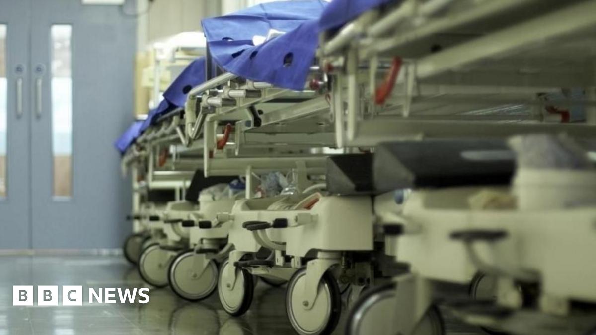 A row of hospital beds are in a corridor in a medical facility they have the blue blankets visible. The picture is taken from an angle on the ground so the wheels of the beds are in focus.