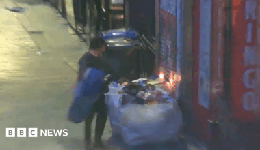 A CCTV image shows Daniel Byrne standing next to a large sack of rubbish in front of the doors of the Beatles Museum. The rubbish is starting to burn, and flames can be seen rising from it.