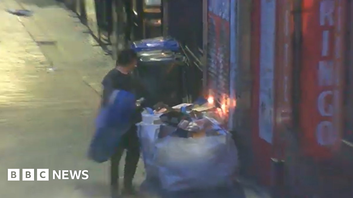 A CCTV image shows Daniel Byrne standing next to a large sack of rubbish in front of the doors of the Beatles Museum. The rubbish is starting to burn, and flames can be seen rising from it.