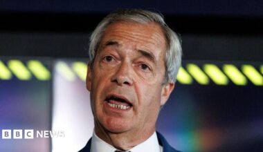 Nigel Farage, wearing a white shirt and navy blazer, tilts his head to one side as he speaks