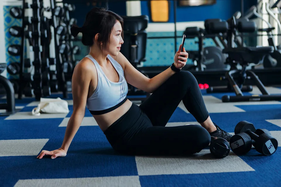 Side view of young athletic Asian woman using exercise tracking app on smartphone to monitor her training progress after exercising at gym. Practice Yoga. Personal workout concept. Online training class. Training app.