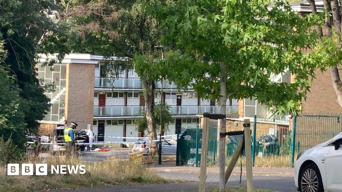 An area in front of a small block of flats is cordoned off with blue and white police tape. There is a police officer in a black hat and fluorescent yellow vest guarding the tape. A forensic officer in a full white suit is looking inside a black car
