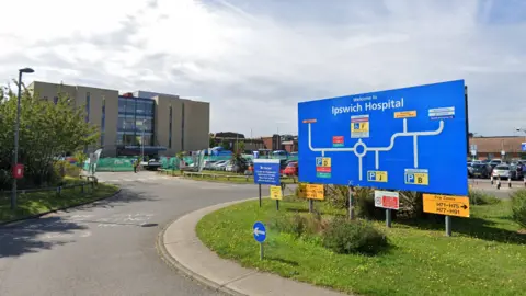 Google The outside of Ipswich Hospital - a large blue sign is in the foreground on a roundabout. The sign has a map of parking locations and information for patients. The hospital building is in the background and has a glass front with brick sides. 