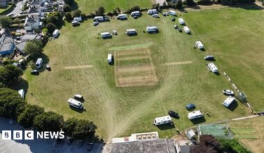 The image shows an aerial view of a sports field with several caravans and vehicles parked around its edges. The central rectangular pitch suggests it's used for cricket. Surrounding the field are residential houses, trees, and a parking area with cars.