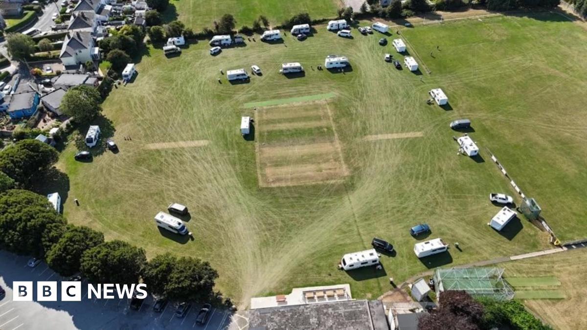 The image shows an aerial view of a sports field with several caravans and vehicles parked around its edges. The central rectangular pitch suggests it's used for cricket. Surrounding the field are residential houses, trees, and a parking area with cars.