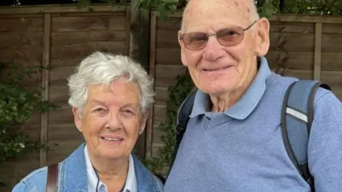 Gemma Dillon/BBC The image shows a couple.
The lady on the left is wearing a denim jacket and has short grey hair. 
The man on the right is wearing a blue jumper with a blue rucksack on his shoulders. he is wearing tinted glasses. 
Both are stood infront of a wooden fence