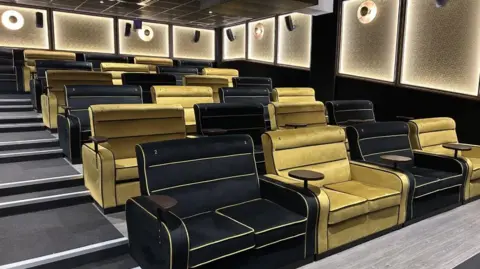 The inside of a cinema with black and gold two-seater sofa-style chairs. Stairs are on the left and there are little tables in between each two-seater. 
