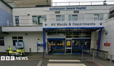 Entrance to hospital, a two-storey white building with white-framed windows. There are four blue doors, with a white one in the centre. A white sign with dark blue writing indicated "All Wards and Departments". There is a grey railing to the right and a motorcycle with rider in yellow hi-vis to the left.