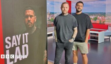 On the left, Danny Tanti, who has short black hair combed forward, and a black beard and moustache. Jonny Human, on the right, has short dark hair combed up on top and cropped short on the sides. He has a light brown full beard and moustache. They are both wearing a Say It Lad branded T- shirt, and are standing inside the BBC Look East studio, with their logo and an image of Jonny on the tower screen.