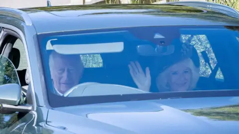 PA Media A smiling King Charles III and Queen Camilla, who is also waving her right hand, as they arrive to attend a Sunday church service at Crathie Kirk, near Balmoral.
The King is driving a dark grey Audi and the Queen is sitting in the front seat.