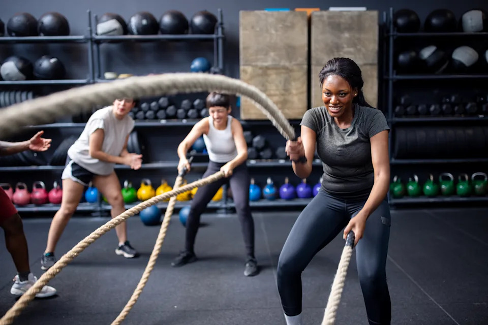 Female fitness enthusiasts engage in a vigorous battle ropes workout, supported by friends cheering them on at a cross-training gym.