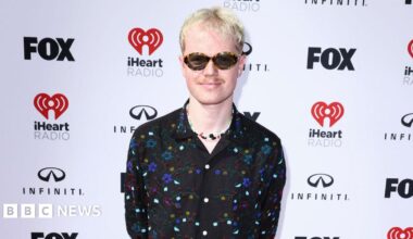 Lloyd Wakefield stands in front of a white backdrop that has Fox, iHeart Radio and Infiniti logos. He has short light blonde hair, a moustache and is wearing sunglasses. He is smiling at the camera and is wearing a black, floral beaded shirt with a colourful charm necklace underneath.