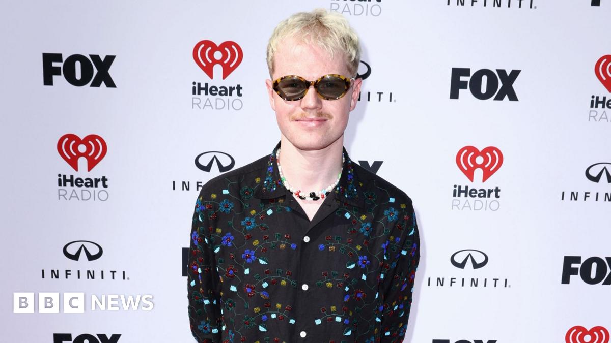 Lloyd Wakefield stands in front of a white backdrop that has Fox, iHeart Radio and Infiniti logos. He has short light blonde hair, a moustache and is wearing sunglasses. He is smiling at the camera and is wearing a black, floral beaded shirt with a colourful charm necklace underneath.