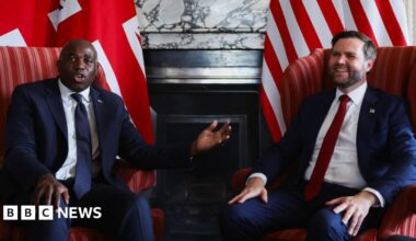 David Lammy, wearing a navy suit and tie, gestures to JD Vance, who is wearing a navy suit and red tie, and has a relaxed smile, as they sit in armchairs during a photo call in front of a British flag and an American flag at Chevening
