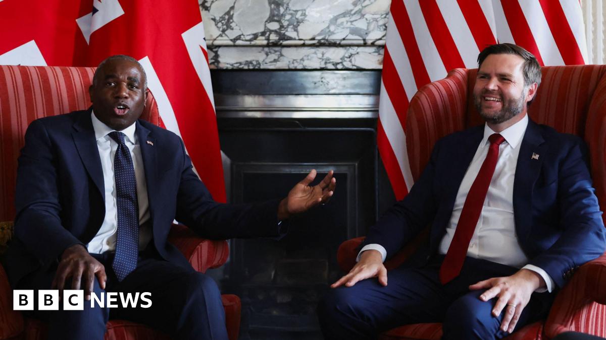 David Lammy, wearing a navy suit and tie, gestures to JD Vance, who is wearing a navy suit and red tie, and has a relaxed smile, as they sit in armchairs during a photo call in front of a British flag and an American flag at Chevening