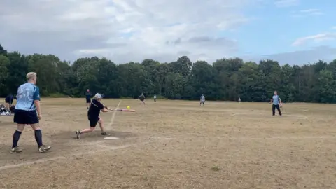 Players from MI6 and the Fuzzy Ducks are mid-game on Wandsworth Common (a dry, grassy field with trees in the background). A female Fuzzy Ducks player wearing a black top, black shorts and black-and-white cap is swinging a bat toward a green-coloured softball. 
Behind her stands Gelske Huyer in a light-blue top and dark shorts in the backstop position. 
To the right of the image stands MI6's male pitcher in a light-blue coloured top and dark trousers. 