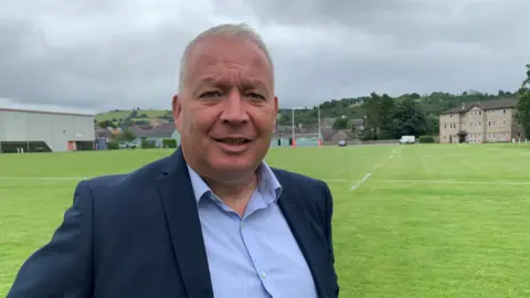 A head and shoulders shot of Dominic on a rugby pitch. He has a blue jacket and a light blue shirt on. He has short grey hair and is smiling at the camera.