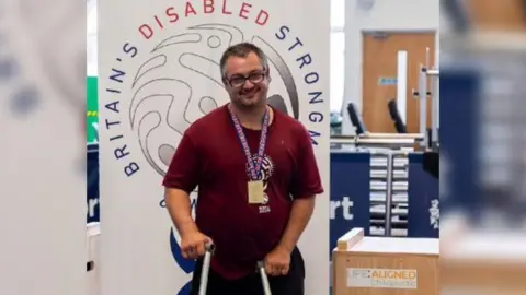 Gary Clarke Gary Clarke wearing a burgundy t shirt and glasses with a purple lanyard. Gary is standing in front of a big sign which reads Britain's Disabled Strongman. He is standing with crutches. 