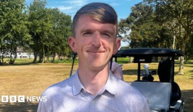 David Gascombe smiles at the camera, he has light brown hair in a side part and is wearing a lilac shirt. There is a golf buggy in the background.