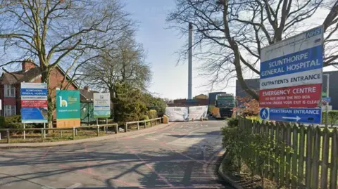 Google The entrance to a hospital with fencing either side of a road going into it. Either side of the fencing you can see signs saying 'NHS Scunthorpe General Hospital, Outpatients Entrance, Emergency Centre'.