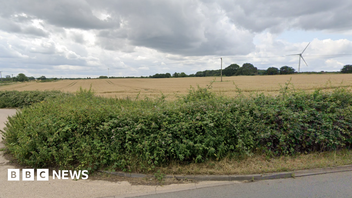 Land off Heath Road in Bagworth, Leicestershire, with shrubs in the forefront at the roadside and trees and a wind turbine in the distance on a dry cloudy day.