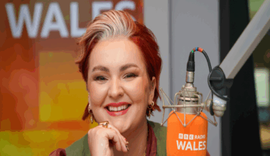 A woman with short died red hair, wearing a red shirt and green waistcoat and smiling at the camera. She has her chin leaning on one hand, and is wearing gold earrings, rings and necklace. In front of her is an orange microphone with BBC Radio Wales written on it.