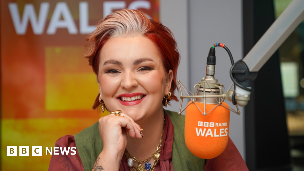 A woman with short died red hair, wearing a red shirt and green waistcoat and smiling at the camera. She has her chin leaning on one hand, and is wearing gold earrings, rings and necklace. In front of her is an orange microphone with BBC Radio Wales written on it.