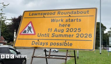 Vehicles approacing the Lawnswood roundabout in Leeds, with a large green road sign on the left, indicating the oncoming roundabout