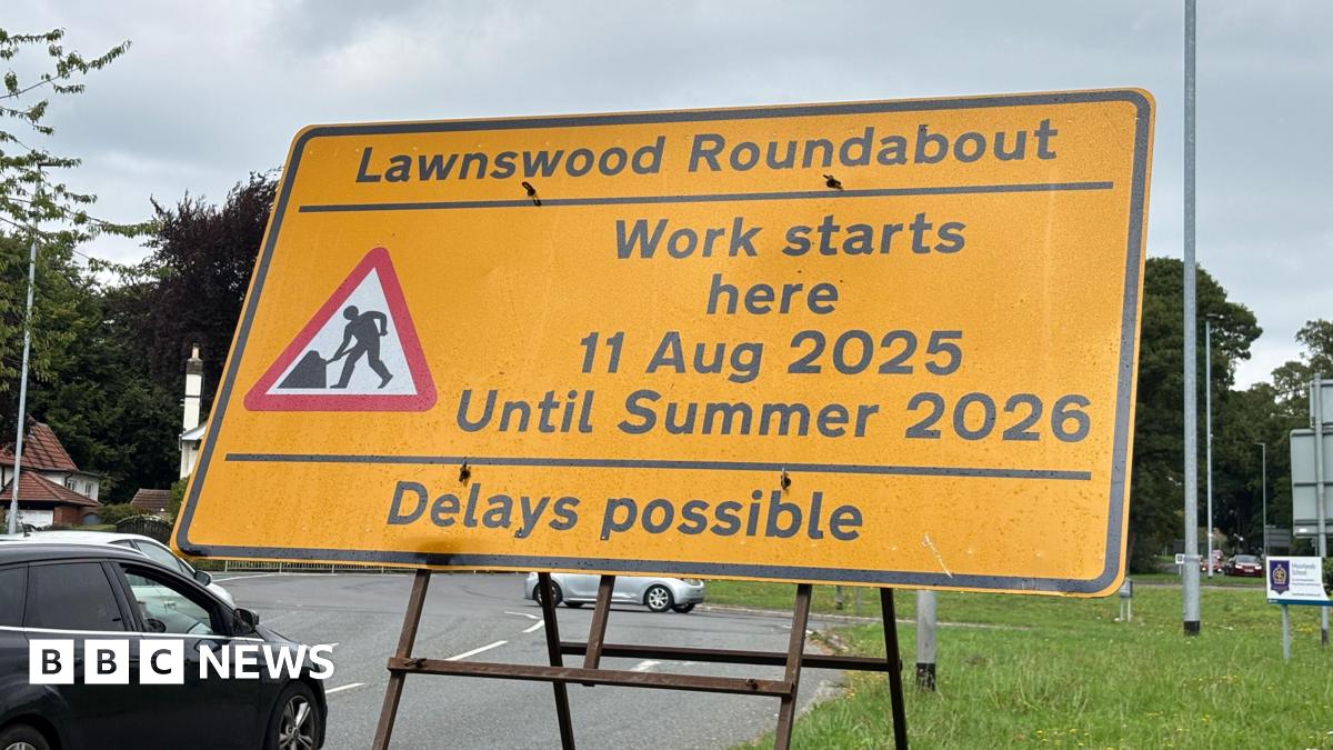 Vehicles approacing the Lawnswood roundabout in Leeds, with a large green road sign on the left, indicating the oncoming roundabout