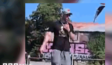 A grainy image of a white man in grey jogging bottoms, black T-shirt and a grey cap. In the background is a building with graffiti on it, surrounded by bushes.