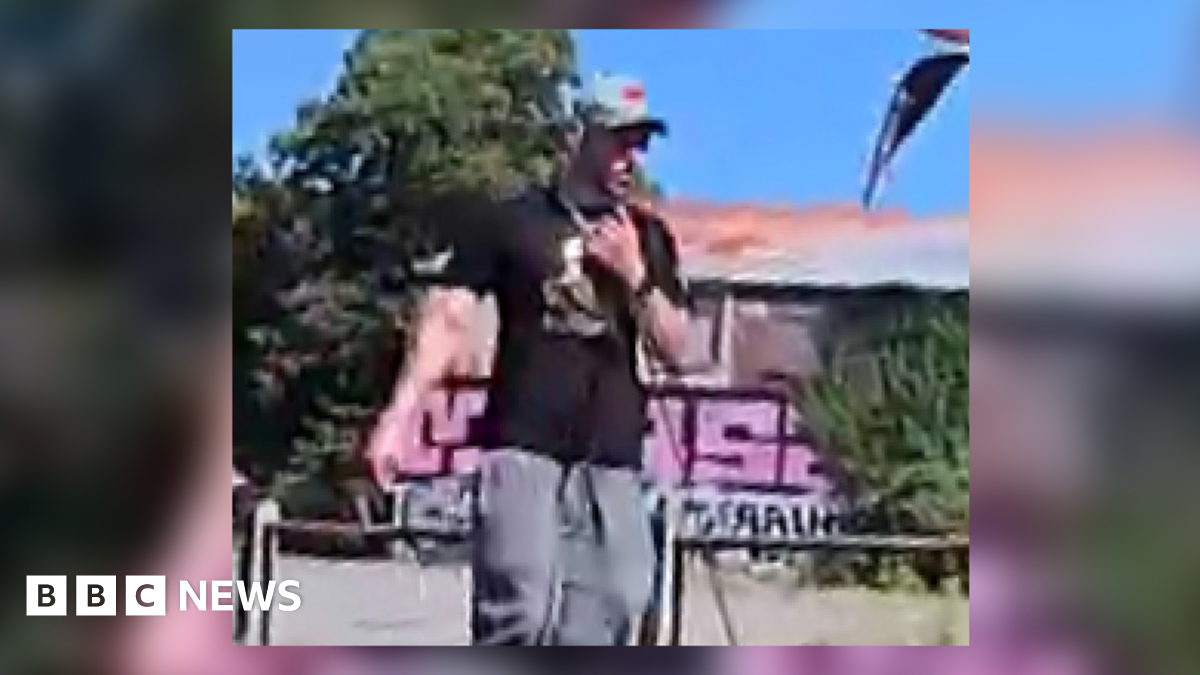 A grainy image of a white man in grey jogging bottoms, black T-shirt and a grey cap. In the background is a building with graffiti on it, surrounded by bushes.