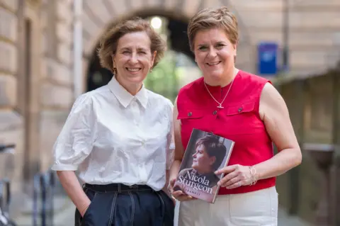 PA Media Kirsty Wark has short brown hair and smiles at the camera. She wears a white shirt and dark trousers. She stands next to Nicola Sturgeon who smiles at the camera. She has short fair hair and wears a red top and white trousers and holds a copy of her book which has her face profile pictured and is called Frankly 