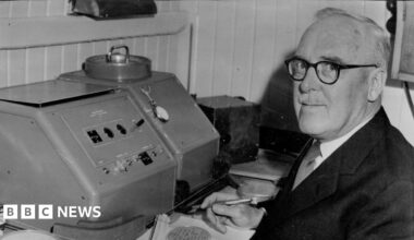 Black and white image of Sir Ernest Marsden, who is wearing a pair of spectacles, looking at the camera as he poses while sitting by a scientific machine.