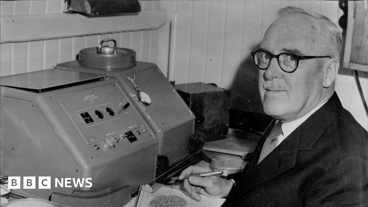 Black and white image of Sir Ernest Marsden, who is wearing a pair of spectacles, looking at the camera as he poses while sitting by a scientific machine.
