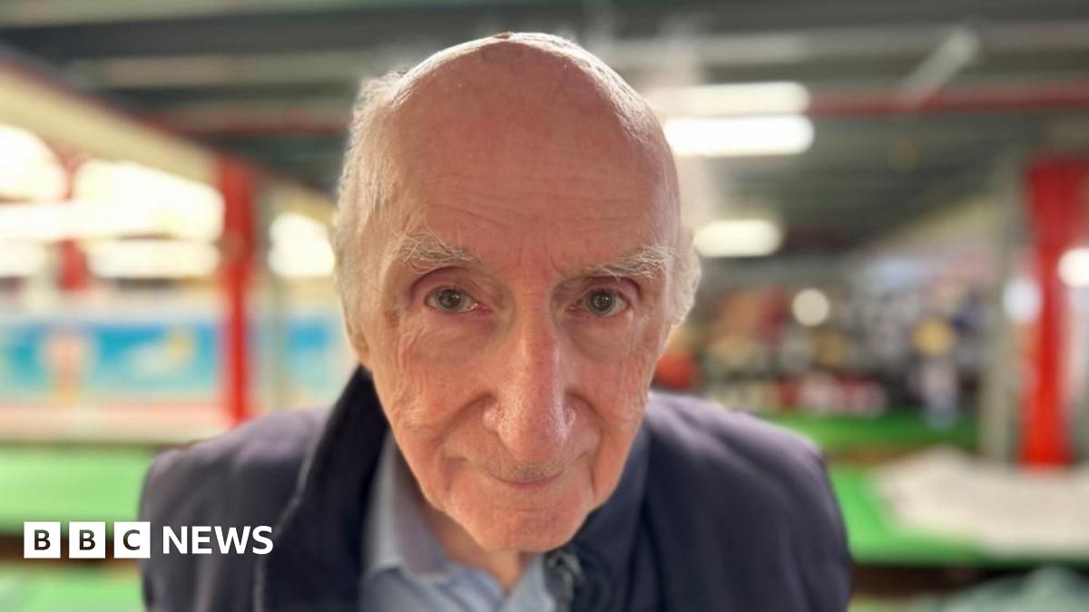 Don Cameron smiles at the camera from the ground floor of his Bedminster balloon factory with cutting tables in the background. He is an elderly man with white hair and blue eyes.