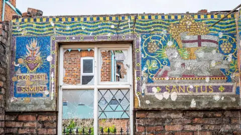 Great Places Housing Group A broad, horizontal mosaic straddles a broken window on a partially demolished wall. The left side of the mosaic shows the Sacred Heart of Jesus, while the other shows a lamb with the flag of St George waving above.