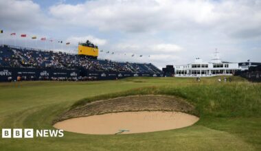 a golf course, with a sandy bunker in the foreground. On the left there is a large stand, approximately half full of fans. Above that, there is a small TV gantry for cameras. To the right of the stand, a large white building is pictured. On the course, there are several golf players and caddies in the distance.